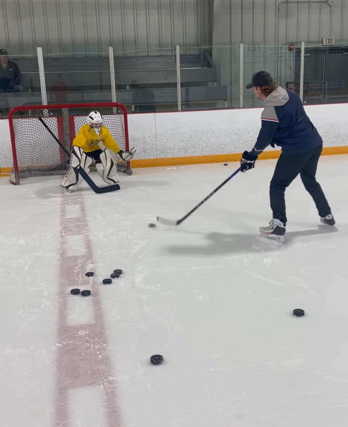goalie - Fresh Legs Hockey School Youth hockey goalie in a yellow jersey crouches in front of the net as a skater in a blue jacket shoots on an indoor rink.