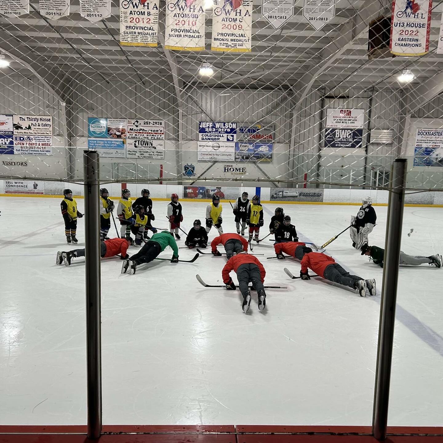 pushups - Fresh Legs Hockey School Youth hockey players gather in a huddle on the ice during practice, with a goalie standing nearby.